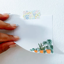 Load image into Gallery viewer, A page of the square white memo pad with the pots of plants on the bottom corner designed is stuck onto a white wall with colorful floral washi tape.  A hand is lifting a left bottom corner of the memo pad revealing the thickness of one sheet of memo pad. 
