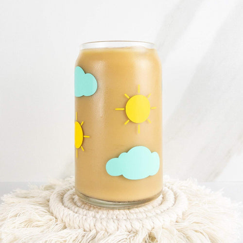 Yellow suns and mint clouds are on this 16 oz can shaped glass cup. It is filled with coffee on a coaster. 
