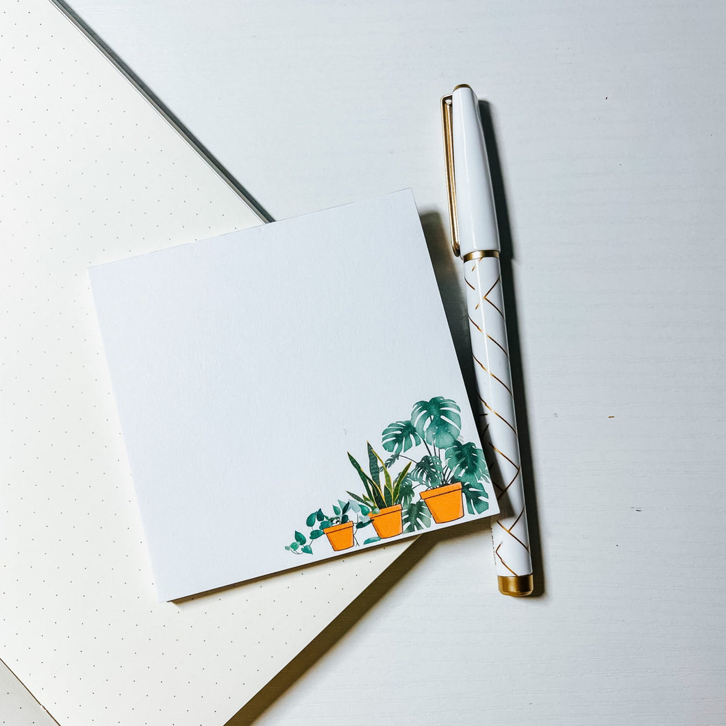 Square white color memo pad with pots of devil ivy, snake and monsterra plant in the right bottom corner is laid out on spreaded bulleet journal next to white and gold patterned pen. 